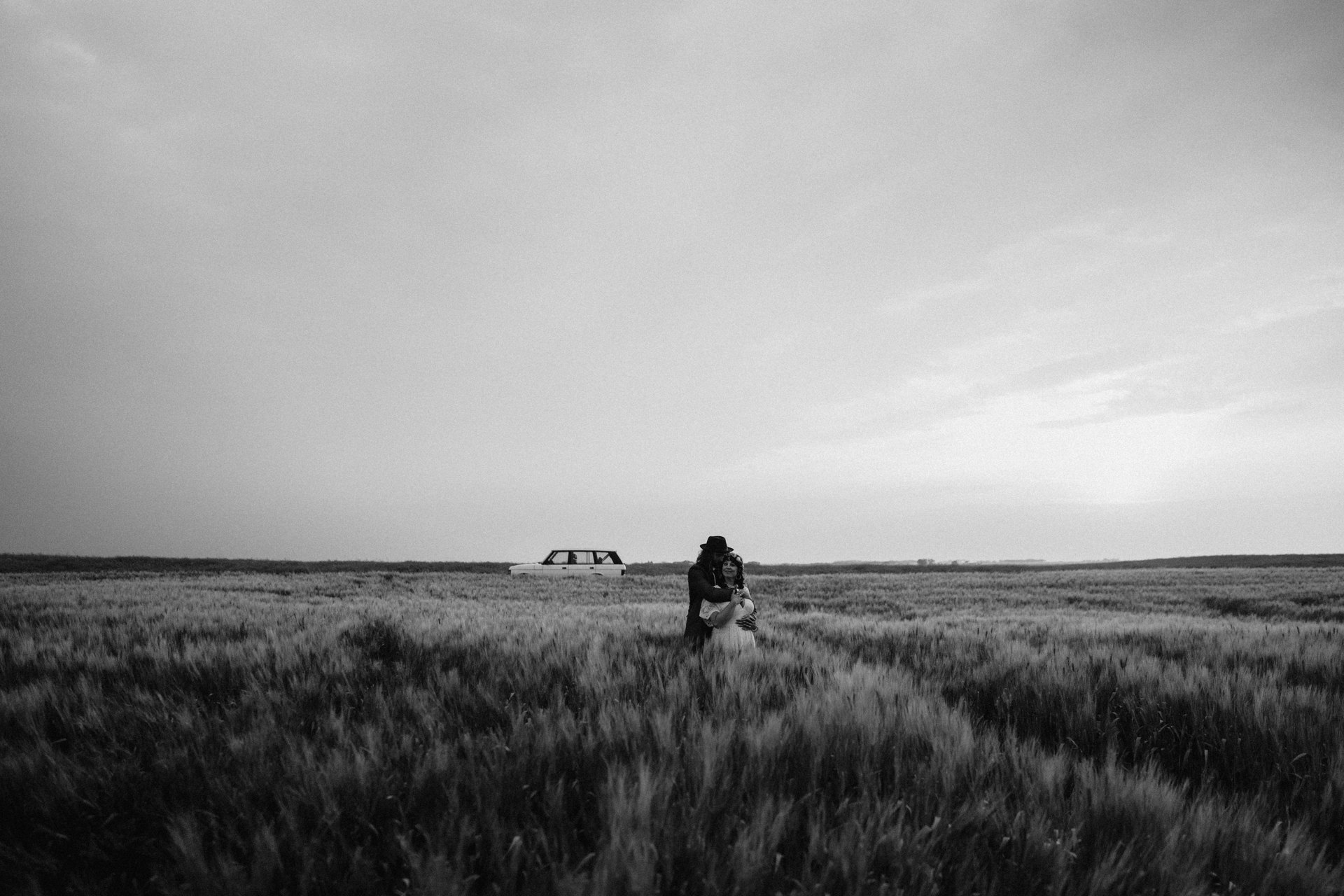 Cayle and Nathaniel holding each other in a wide wheat field under a pale sky, an old station wagon parked on the horizon