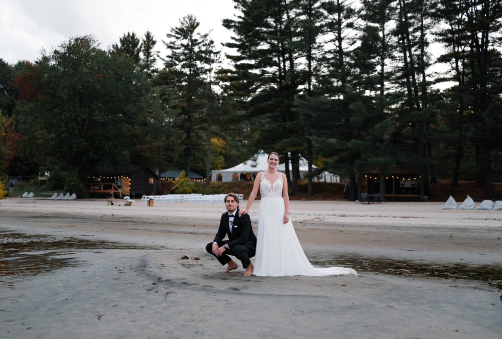 Paige kneeling barefoot beside Greg on the sand at Anupaya Cabin Co., tall pines behind